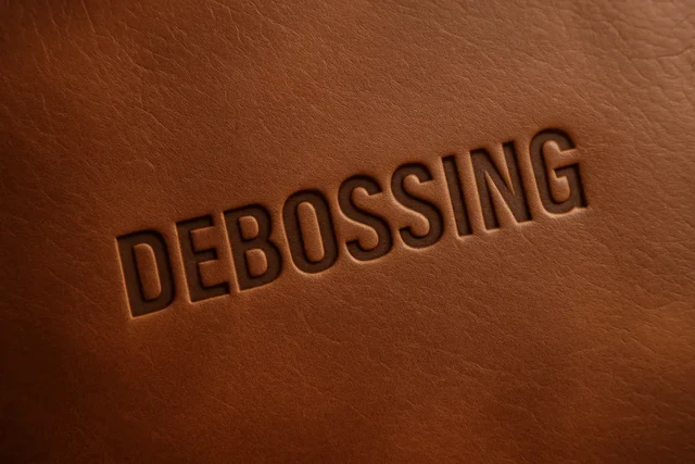 Close-up of the word "DEBOSSING" recessed into textured brown leather, showing detailed debossed lettering and rich leather grain